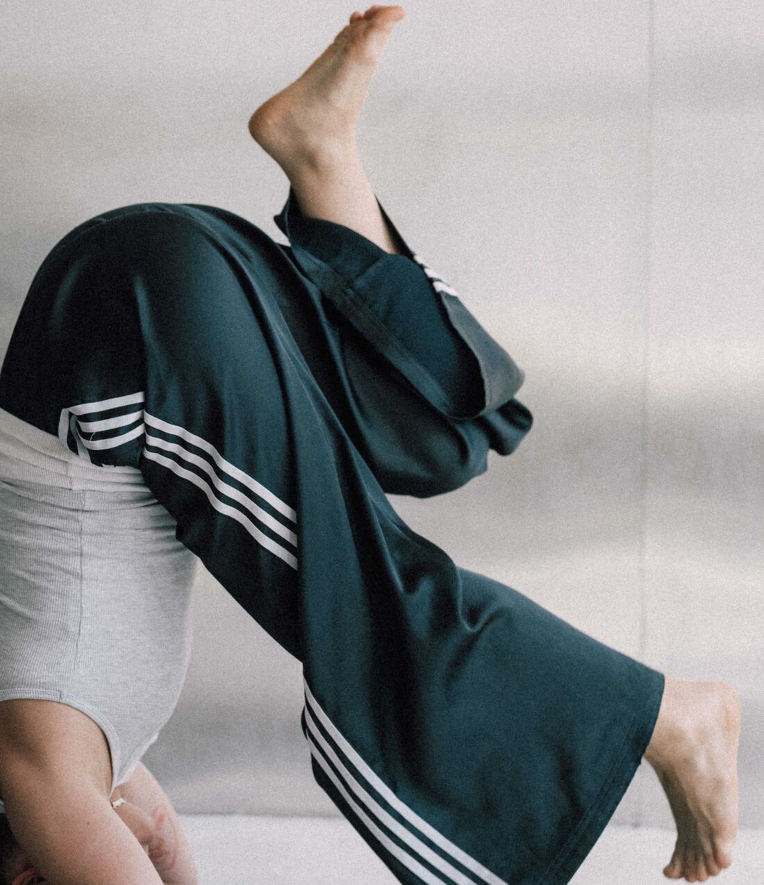 Someone upside down in a shoulderstand, bare feet in the air, in a deep green striped tracksuit against a pale wall.