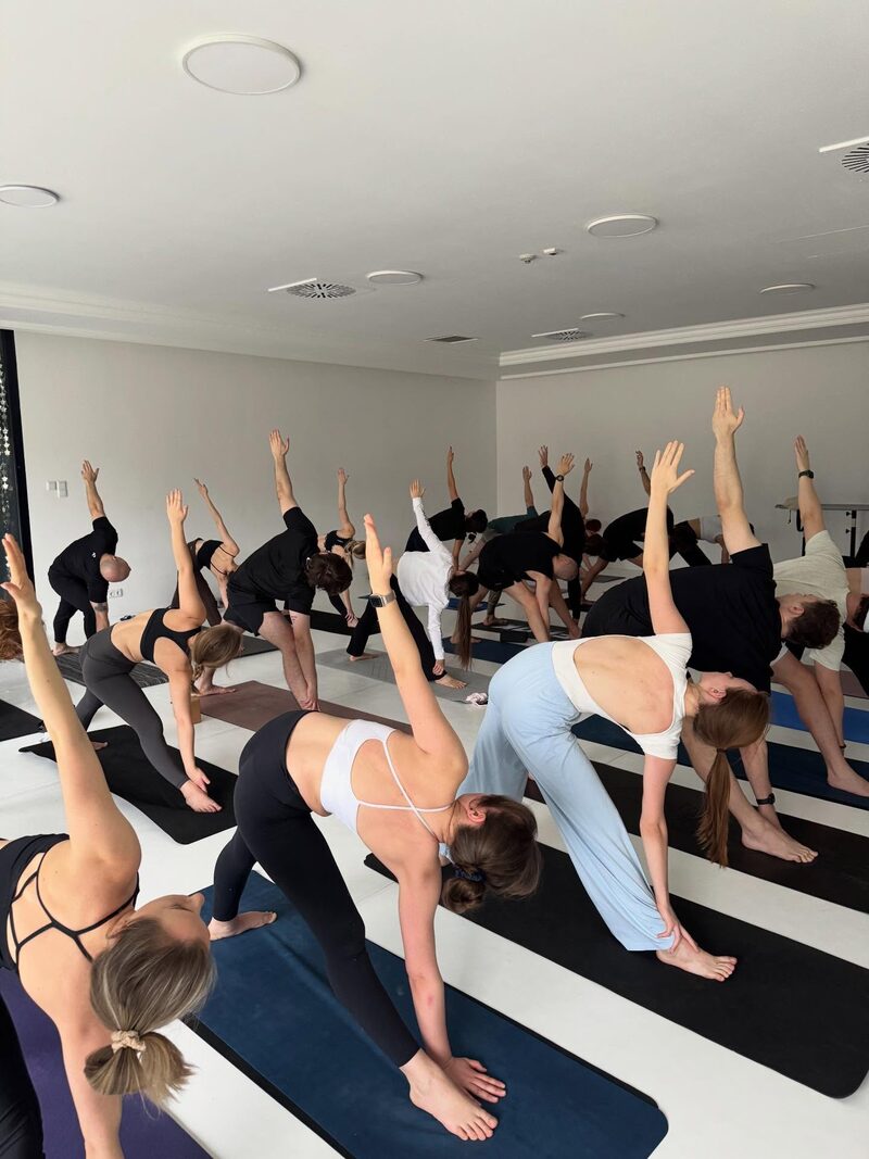 A full studio in triangle pose, two dozen people turned away from the camera, one arm raised.